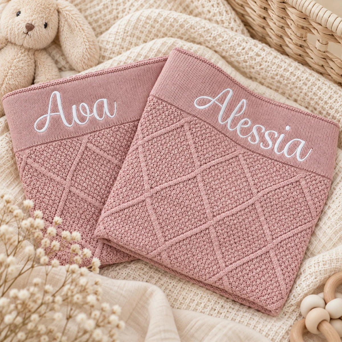 Two dusty pink knitted baby blankets white embroidered names 'Ava' and 'Alessia' on the waistbands, styled on a beige textured blanket with a bunny toy, dried flowers, and a wicker basket.