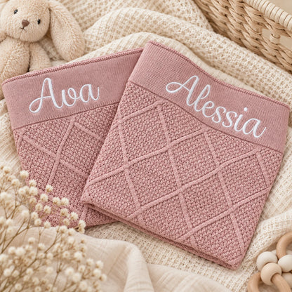 Two dusty pink knitted baby blankets white embroidered names 'Ava' and 'Alessia' on the waistbands, styled on a beige textured blanket with a bunny toy, dried flowers, and a wicker basket.