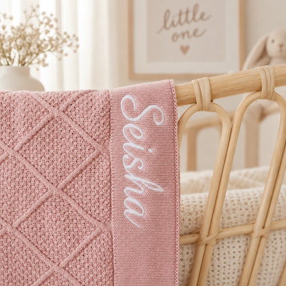 A close-up of a dusty pink knitted baby blanket with a diamond quilted pattern and white embroidered script reading 'Seisha', draped over a light wood rattan crib in a softly lit nursery.