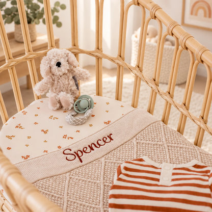 A personalised baby blanket with the name 'Spencer' embroidered in brown, laid inside a light wood rattan crib. The blanket features a cream base with tiny orange and yellow floral prints on the top section and a textured beige diamond-knit pattern on the bottom.
