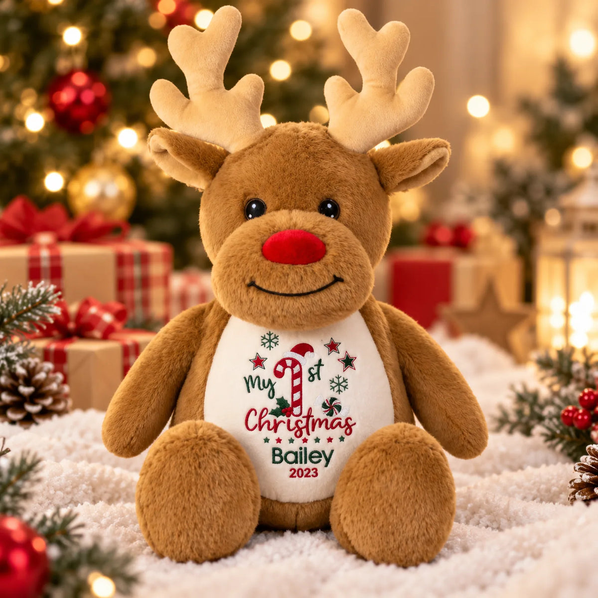 A brown plush reindeer toy sits on a white faux-snow surface with a decorated Christmas tree, wrapped gifts, and warm string lights in the background. The reindeer has a white belly print that reads 'My 1st Christmas Bailey 2023' with a candy cane and snowflake design.
