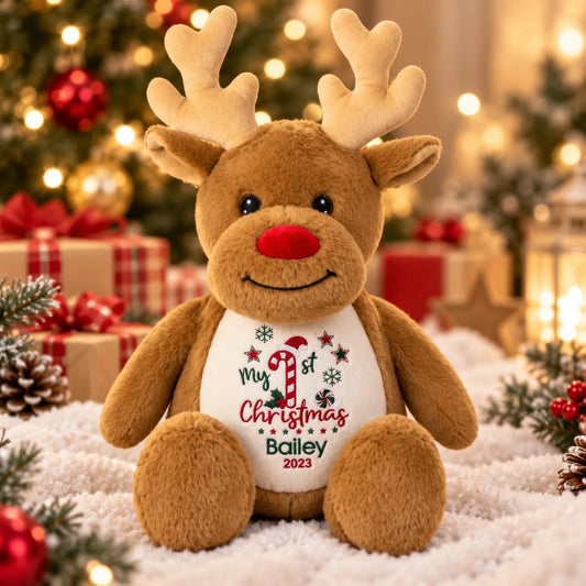 A brown plush reindeer toy sits on a white faux-snow surface with a decorated Christmas tree, wrapped gifts, and warm string lights in the background. The reindeer has a white belly print that reads 'My 1st Christmas Bailey 2023' with a candy cane and snowflake design.