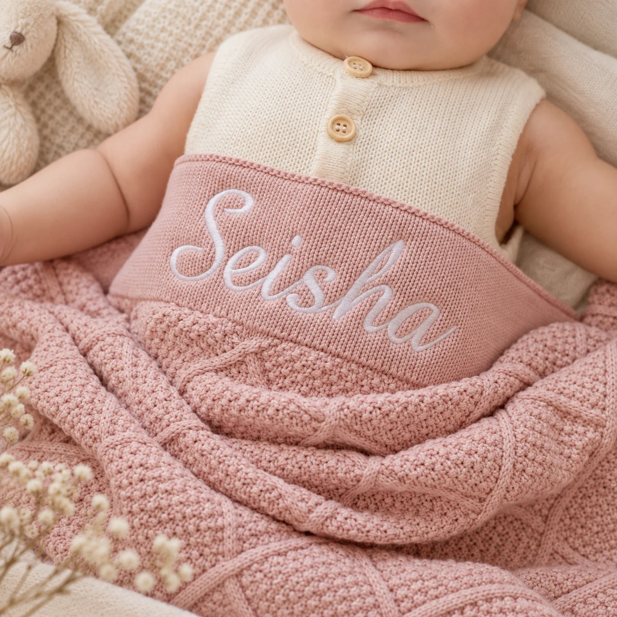 A close-up of a baby doll or infant wearing a cream knit sleeveless top with two wooden buttons and wrapped in a dusty pink cable-knit blanket. The blanket has the name 'Seisha' embroidered in white script across the chest.