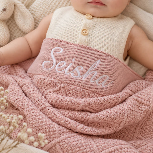 A close-up of a baby doll or infant wearing a cream knit sleeveless top with two wooden buttons and wrapped in a dusty pink cable-knit blanket. The blanket has the name 'Seisha' embroidered in white script across the chest.