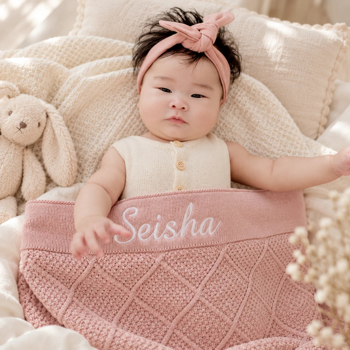 A baby lying on a soft cream blanket with a pink knitted blanket underneath, wearing a pink bow headband and a cream sleeveless top. The blanket is personalized with the name 'Seisha' embroidered in white.