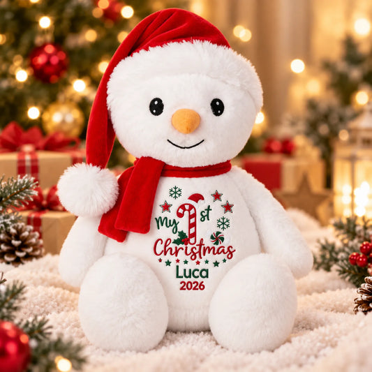 A plush snowman toy wearing a red Santa hat and red scarf, sitting on a white fluffy surface with a festive Christmas background. The front has embroidered text reading 'My 1st Christmas Luca 2026' with a candy cane and snowflake design.