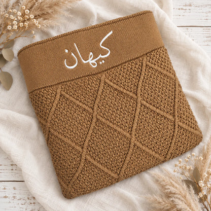 A brown knit pouch or sleeve with a wide elastic band featuring white Arabic script, laid flat on a white fabric surface with dried floral props.