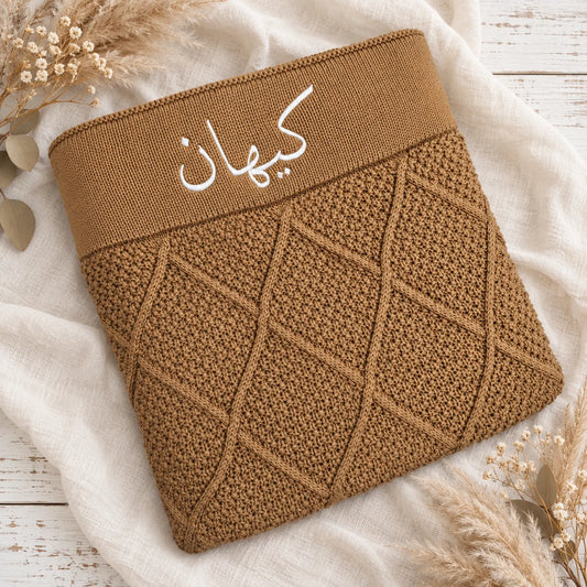 A brown knit pouch or sleeve with a wide elastic band featuring white Arabic script, laid flat on a white fabric surface with dried floral props.