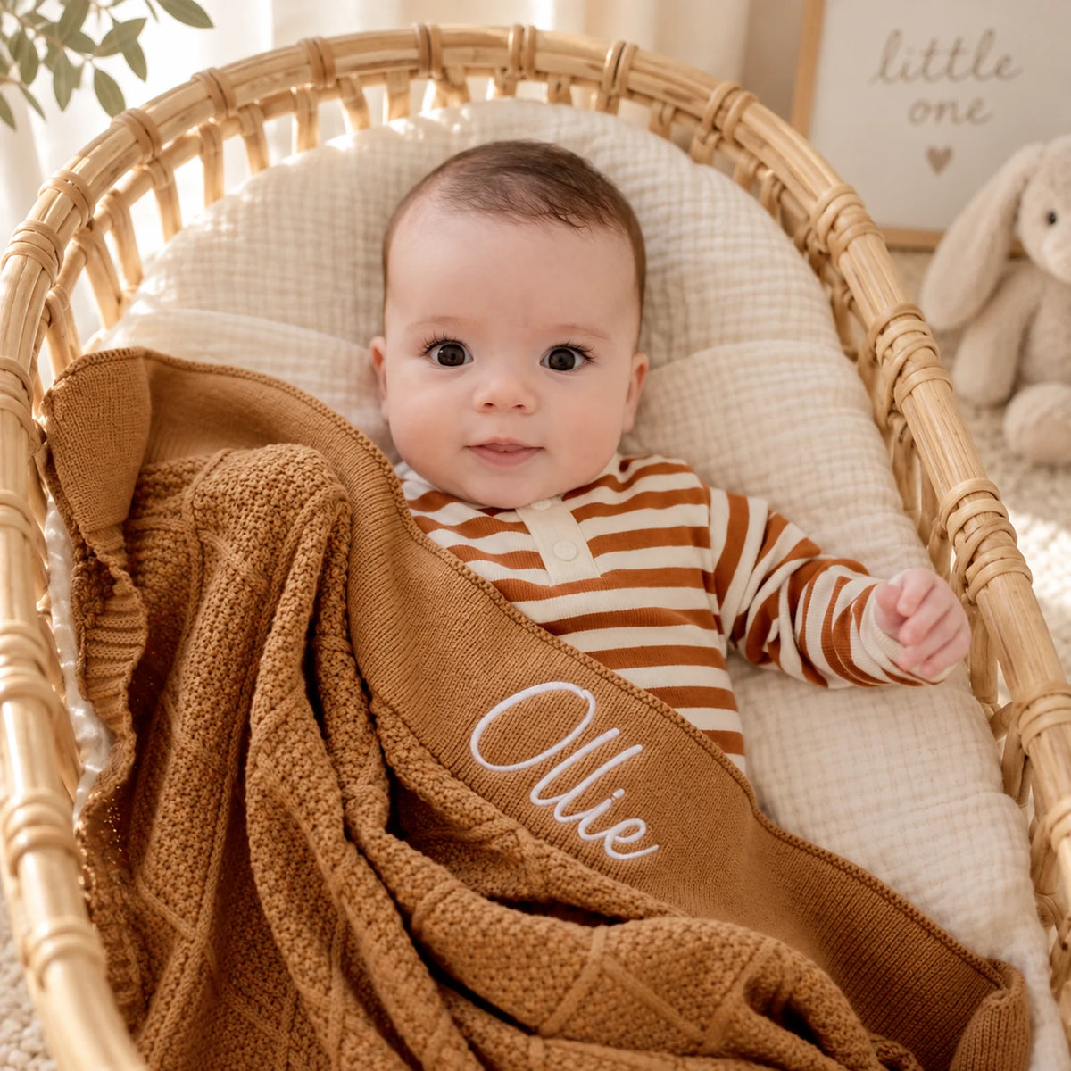 A baby wearing a brown and cream striped long-sleeve bodysuit lies in a woven rattan bassinet, wrapped in a brown knit blanket with the name 'Ollie' embroidered in white. A soft beige wall and a plush bunny toy are visible in the background.