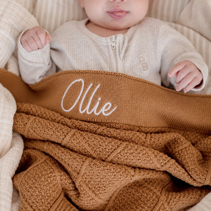 A close-up lifestyle shot of a baby wearing a cream-colored zip-up bodysuit and lying under a personalized brown knit blanket with the name 'Ollie' embroidered in white script.