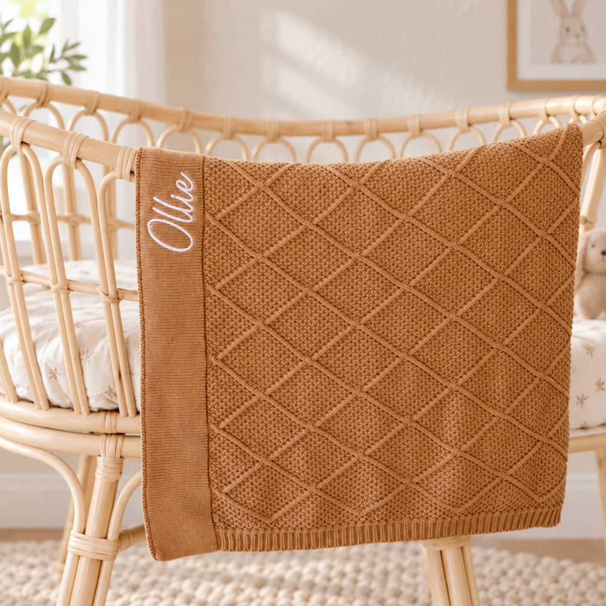 A tan brown diamond knit blankets embroidered with the name Ollie draped over a cot
